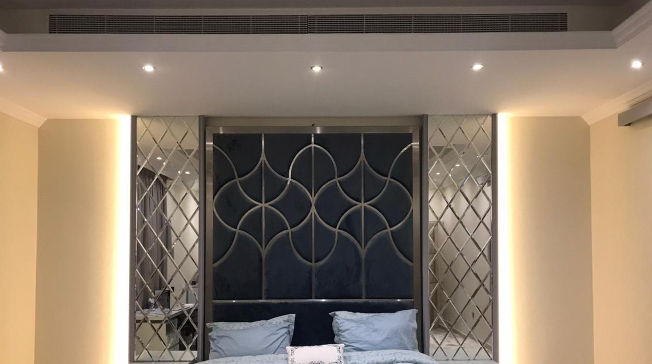 Villa master bedroom with an upholstered headboard wall inlaid with steel arches, flanked by bevelled mirror panels and cove lighting